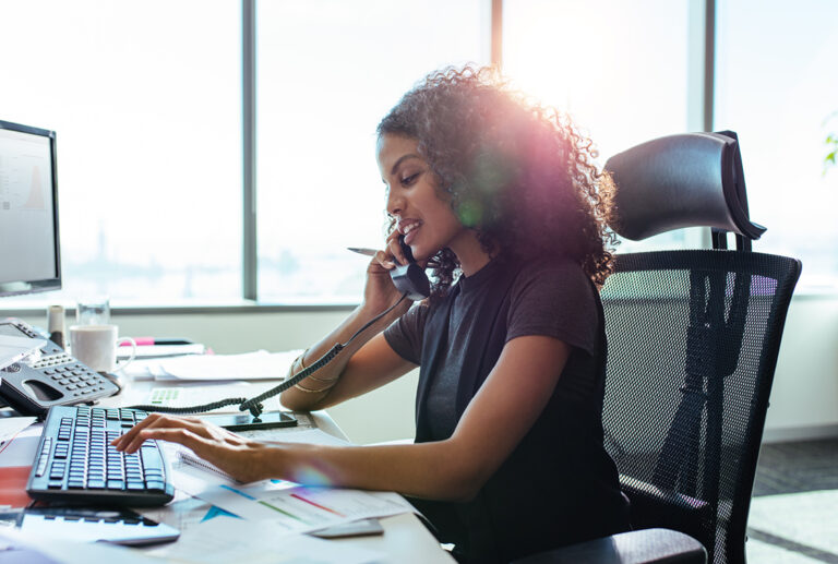 Casually dressed businesswoman with dark brown tightly curled haired talking on the phone at her desk in office.