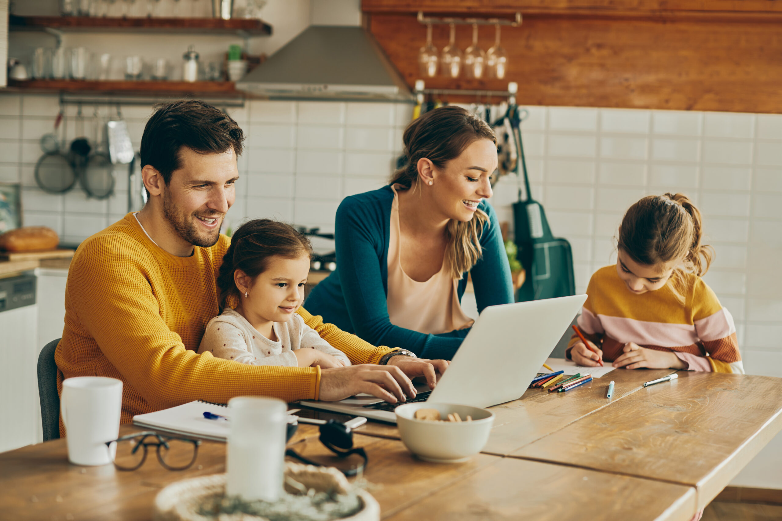 A man and woman with two daughters in a kitchen. One of daughters and father are using laptop while mother and other daughter are colouring on the paper.